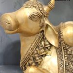 nandi 3 - thebrasshandicrafts.com Gold brass statue of a decorated Nandi bull with ornate jewelry and detailed carvings, shown in profile on a gray background. thebrasshandicrafts.com