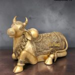 Gold-toned brass statue of a decorative bull with intricate patterns, resting on a wooden surface with a gray background.  thebrasshandicrafts.com