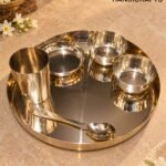 Polished stainless steel serving set on a circular tray with small bowls and a spoon, placed on a beige stone countertop; blurred flowers and brass decor in the background with a 'The Brass Handicrafts' logo in the top right.  thebrasshandicrafts.com