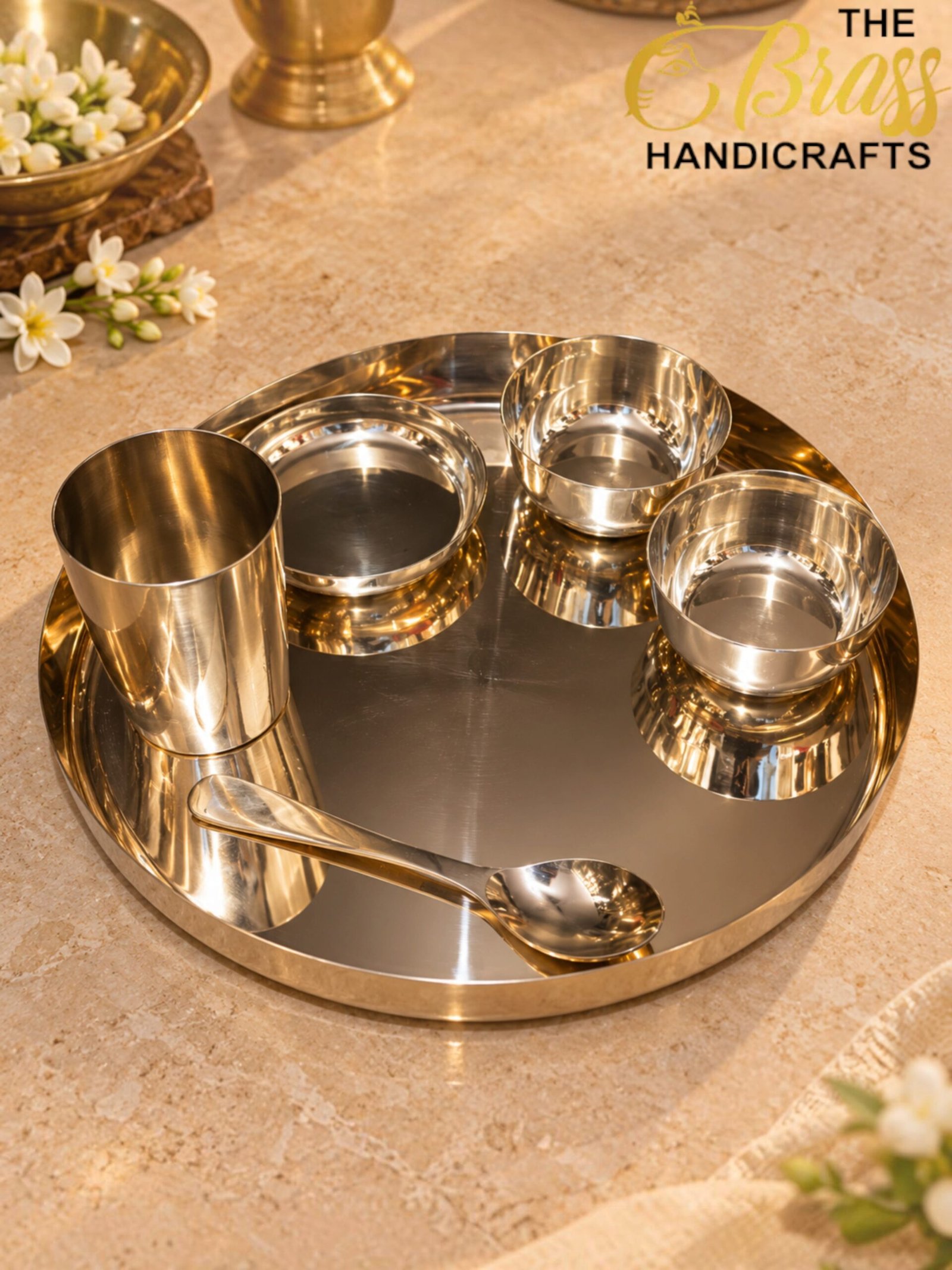 Polished stainless steel serving set on a circular tray with small bowls and a spoon, placed on a beige stone countertop; blurred flowers and brass decor in the background with a 'The Brass Handicrafts' logo in the top right.  thebrasshandicrafts.com