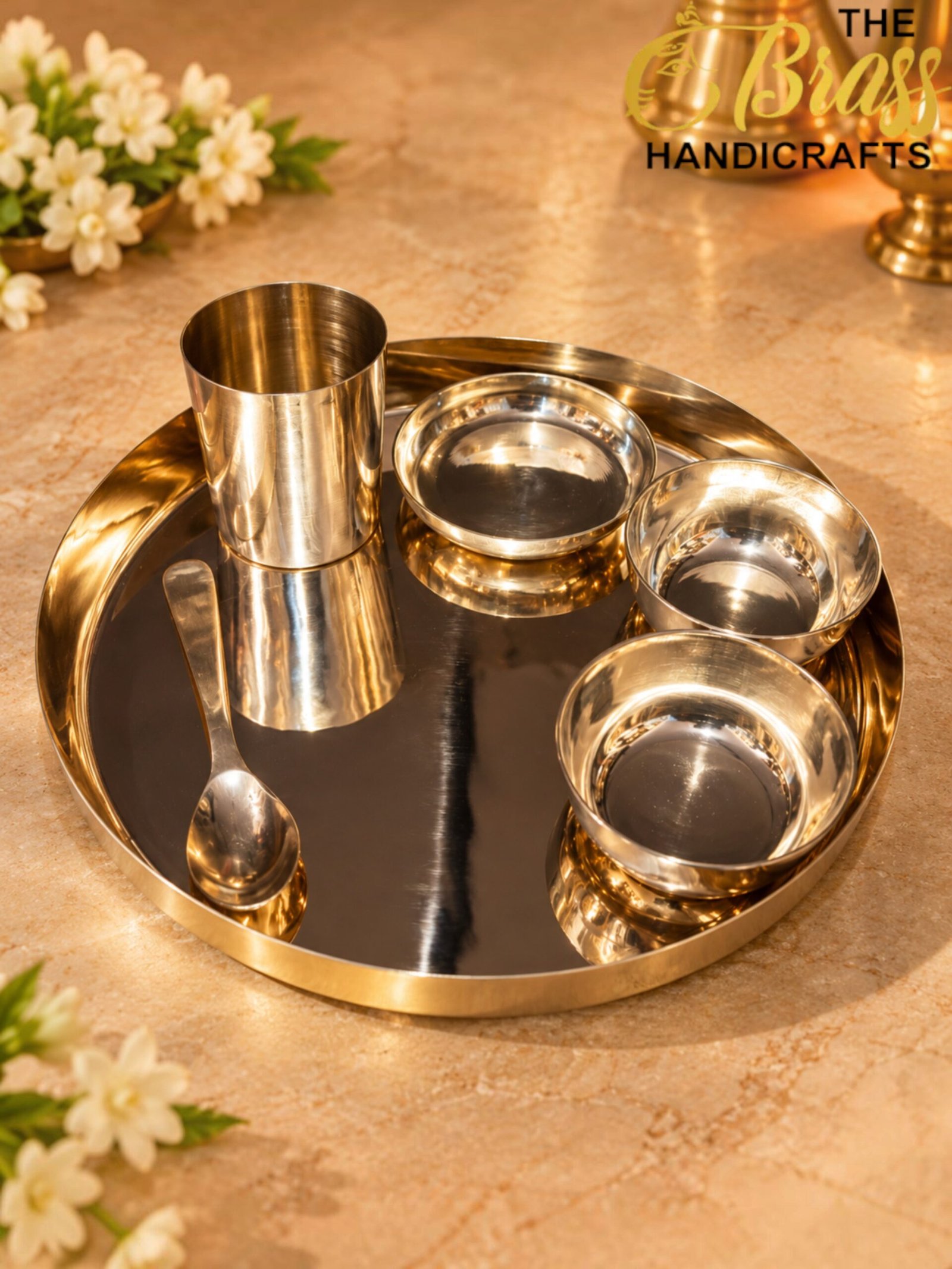 Shiny stainless steel serving set with cups and bowls on a round reflective tray, on a beige countertop with flowers in the background.  thebrasshandicrafts.com