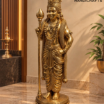 Golden Murugan Shrine in Warm Light 1 - thebrasshandicrafts.com Golden ornate statue of a Hindu deity with a tall crown, layered jewelry, and a scepter, standing on a decorative pedestal in an indoor display space. thebrasshandicrafts.com