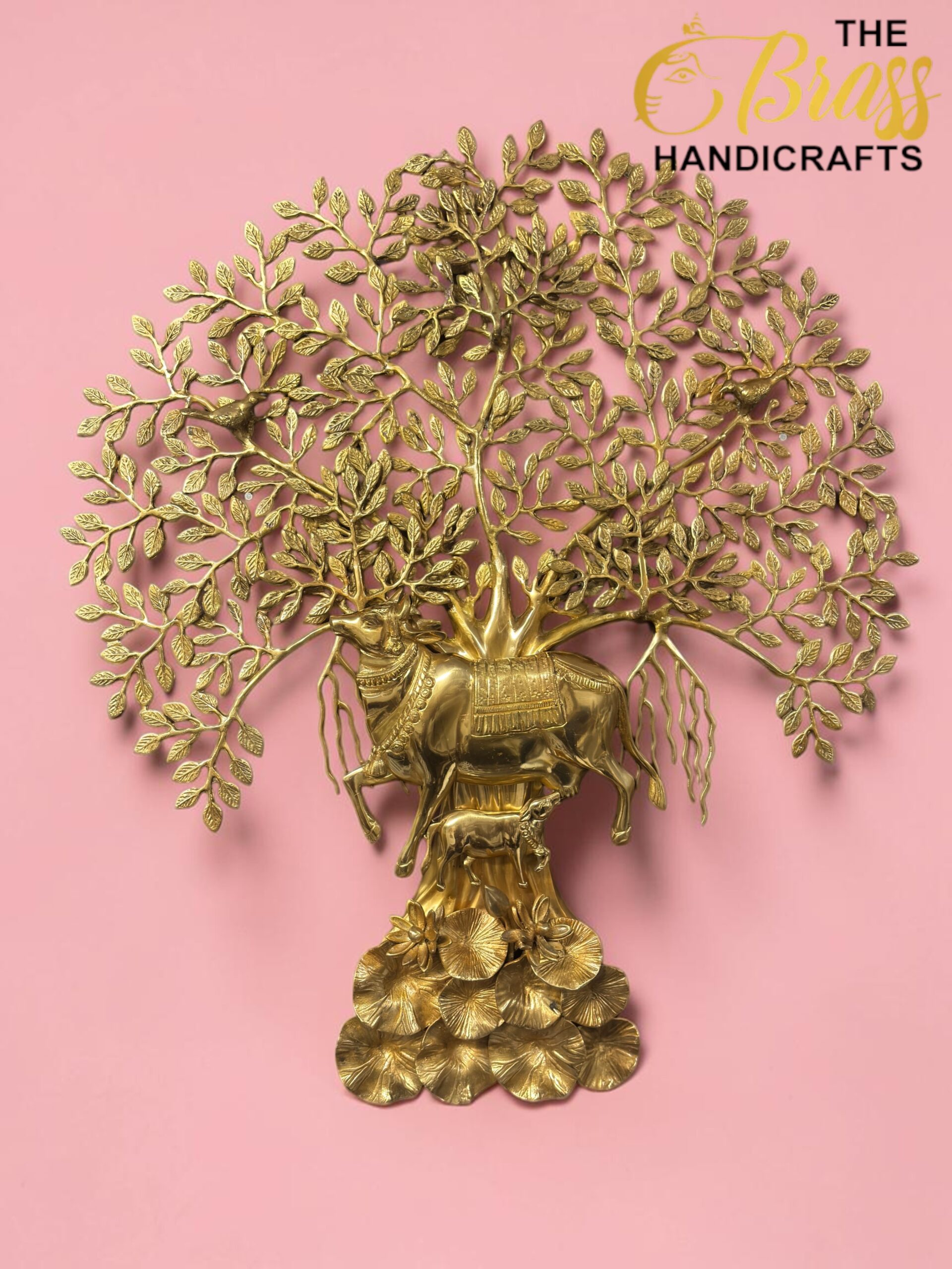 kamdhenu tree 13 - thebrasshandicrafts.com Gold-toned brass tree sculpture with dense leaves on a pink background, featuring a decorative base of circular discs; branding reads THE BRASS HANDICRAFTS. thebrasshandicrafts.com